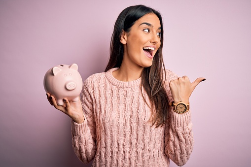 Young beautiful brunette woman holding piggy bank saving money for retirement pointing and showing with thumb up to the side with happy face smiling