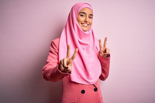Young beautiful brunette businesswoman wearing pink muslim hijab and business jacket smiling looking to the camera showing fingers doing victory sign. Number two.
