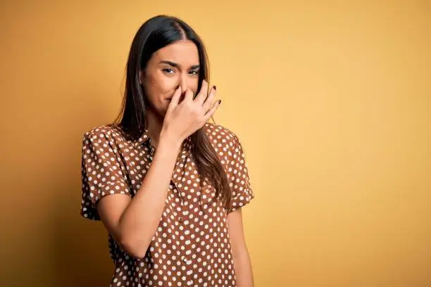Young beautiful brunette woman wearing casual shirt over isolated yellow background smelling something stinky and disgusting, intolerable smell, holding breath with fingers on nose. Bad smell