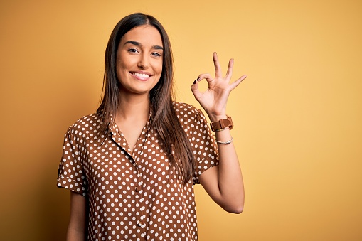 Joven hermosa mujer morena con camisa casual sobre fondo amarillo aislado sonriendo positivo haciendo ok signo con la mano y los dedos. Expresión exitosa. photo