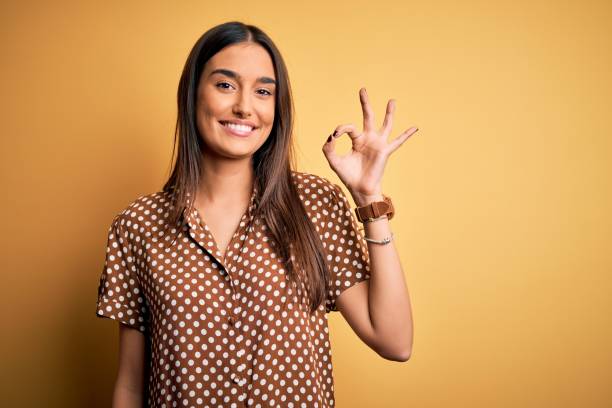jeune belle femme brune utilisant la chemise occasionnelle au-dessus de fond jaune isolé souriant positif faisant le signe d’ok avec la main et les doigts. expression réussie. - doigts en forme de o photos et images de collection
