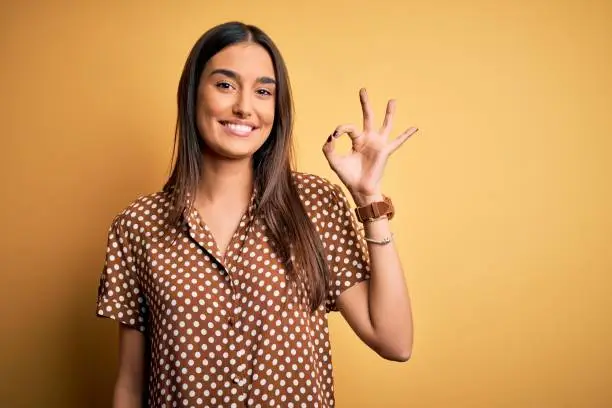 Young beautiful brunette woman wearing casual shirt over isolated yellow background smiling positive doing ok sign with hand and fingers. Successful expression.