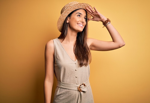 Young beautiful brunette woman on vacation wearing casual dress and hat very happy and smiling looking far away with hand over head. Searching concept.
