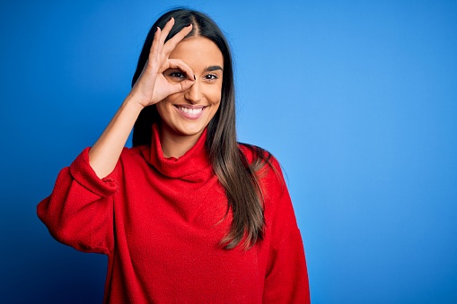 Young beautiful brunette woman wearing red casual sweater over isolated blue background doing ok gesture with hand smiling, eye looking through fingers with happy face.