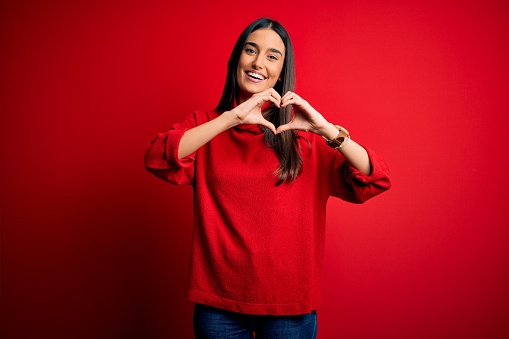 Young beautiful brunette woman wearing casual sweater over isolated red background smiling in love doing heart symbol shape with hands. Romantic concept.