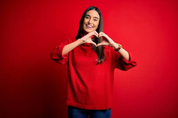 Young beautiful brunette woman wearing casual sweater over isolated red background smiling in love doing heart symbol shape with hands. Romantic concept.