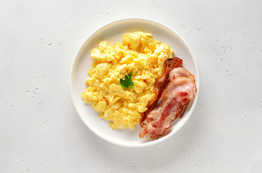 Scrambled eggs and fried bacon on plate over white stone background. Top view, flat lay