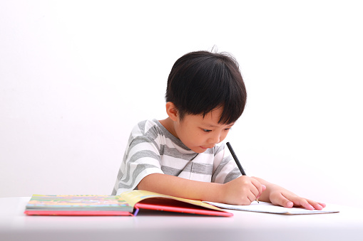 Portrait of little Asian boy sitting at his desk doing homework against white background.