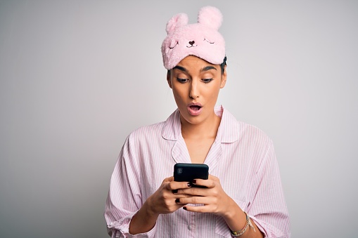 Young beautiful woman wearing pajama and sleep mask having conversation using smartphone scared in shock with a surprise face, afraid and excited with fear expression