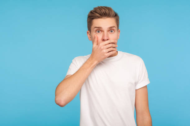 I won't tell anyone! Portrait of scared man in white t-shirt covering mouth with hand to keep silent I won't tell anyone! Portrait of scared man in white t-shirt covering mouth with hand to keep silent, afraid to say secret, looking with intimidated expression. indoor studio shot blue background closed mouth stock pictures, royalty-free photos & images
