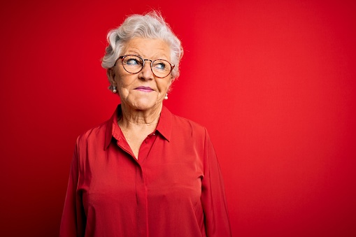 Senior beautiful grey-haired woman wearing casual shirt and glasses over red background smiling looking to the side and staring away thinking.