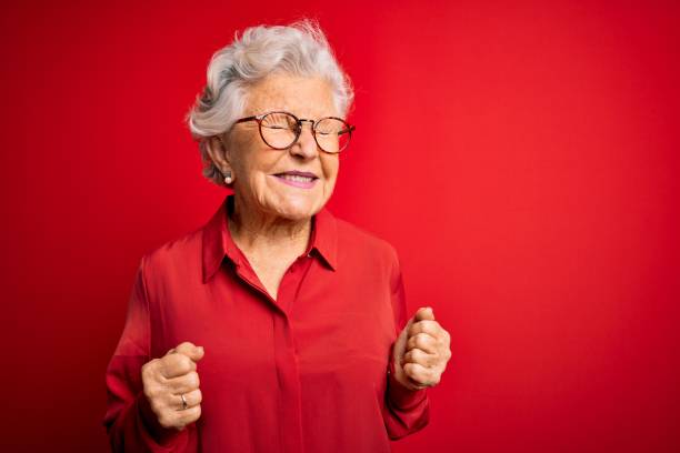 senior beautiful grey-haired woman wearing casual shirt and glasses over red background excited for success with arms raised and eyes closed celebrating victory smiling. winner concept. - women standing fist success imagens e fotografias de stock