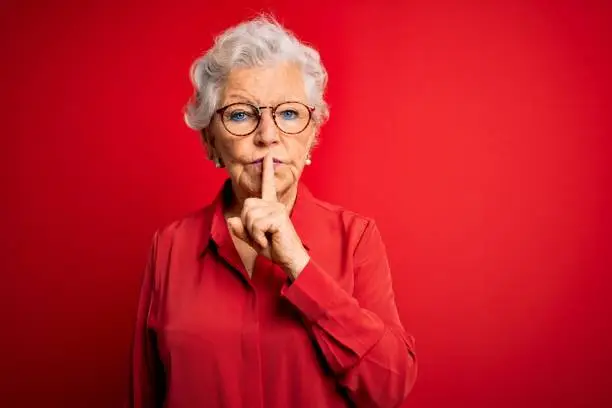 Photo of Senior beautiful grey-haired woman wearing casual shirt and glasses over red background asking to be quiet with finger on lips. Silence and secret concept.