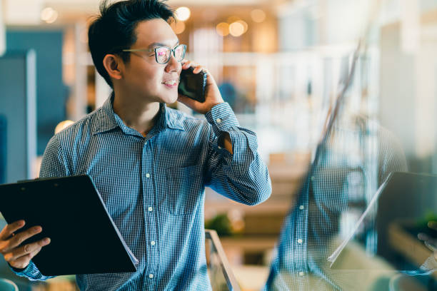 スマート自信アジアのスタートアップ起業家ビジネスオーナービジネスマン笑顔の手は、オフィスの背景に目を引くスマートフォンを使用 - 企業 ストックフォトと画像