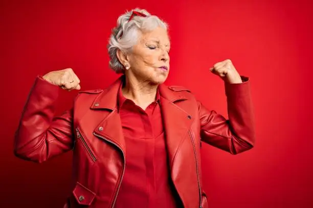 Photo of Senior beautiful grey-haired woman wearing casual red jacket and sunglasses showing arms muscles smiling proud. Fitness concept.