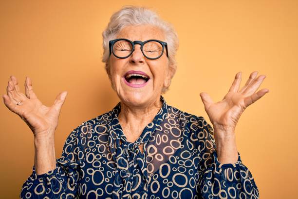 старшая красивая седовласая женщина в непринужденной рубашке и очках на желтом фоне празднует безумный и сумасшедший за успех с поднятыми  - cheerful women shouting happiness стоковые фото и изображе�ния