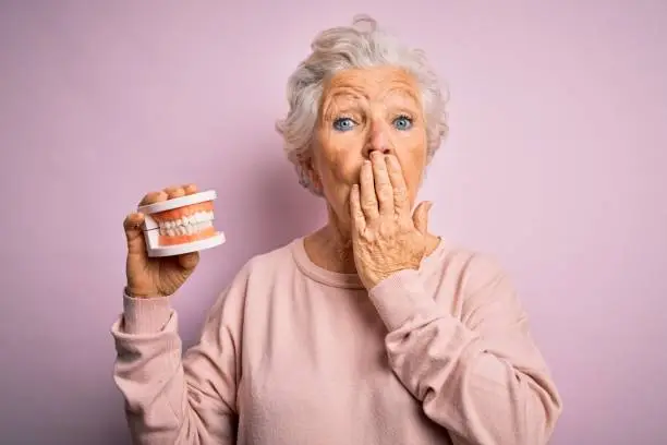 Senior beautiful grey-haired woman holding plastic denture teeth over pink background cover mouth with hand shocked with shame for mistake, expression of fear, scared in silence, secret concept