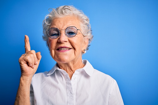Senior beautiful woman wearing elegant shirt and glasses over isolated blue background pointing finger up with successful idea. Exited and happy. Number one.