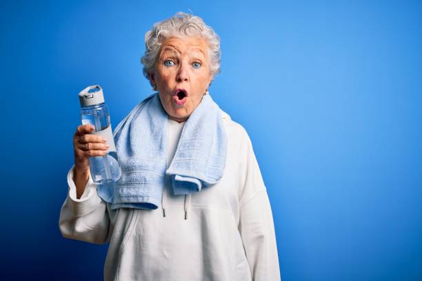 старшая красивая спортивная женщина, держащая бутылку воды, стоящую над изолированным синим фоном, напугана в шоке от неожиданного лица, бо - portrait senior adult confusion women стоковые фото и изображения
