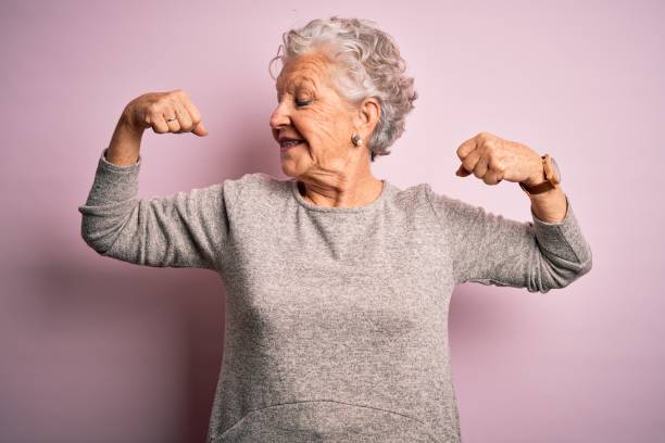 bella donna anziana che indossa una maglietta casual in piedi su uno sfondo rosa isolato che mostra i muscoli delle braccia sorridenti orgogliosi. concetto di fitness. - muscoli foto e immagini stock