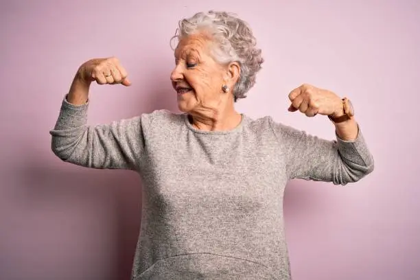 Photo of Senior beautiful woman wearing casual t-shirt standing over isolated pink background showing arms muscles smiling proud. Fitness concept.