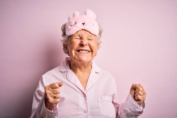 mujer hermosa senior con máscara de sueño y pijama sobre fondo rosa aislado excitado para el éxito con los brazos levantados y los ojos cerrados celebrando la victoria sonriendo. concepto ganador. - máscara para los ojos fotografías e imágenes de stock