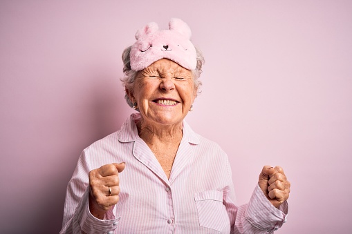Senior beautiful woman wearing sleep mask and pajama over isolated pink background excited for success with arms raised and eyes closed celebrating victory smiling. Winner concept.
