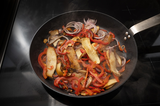 Steaming cooking pan with roasting vegetables like king oyster mushrooms, red peppers and onions on a black stove, copy space, selected focus, narrow depth of field