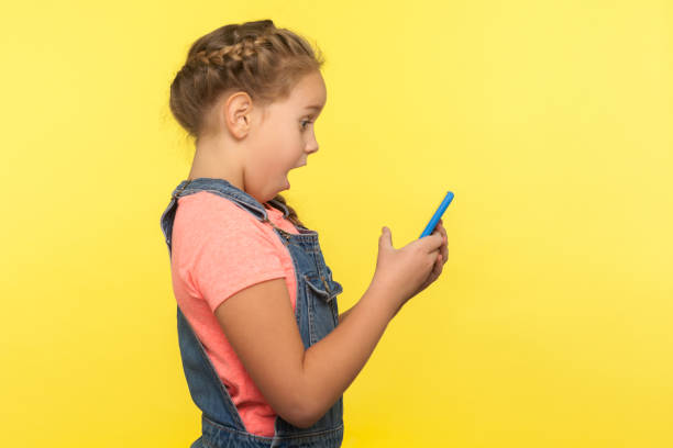 боковой вид шокированной маленькой девочки, читающих удивительное сообщение на мобильный телефон, в чате в социальной сети - surprise child little girls shock стоковые фото и изображения