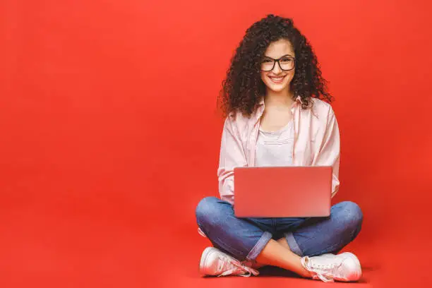 Photo of Happy young curly beautiful woman sitting on the floor with crossed legs and using laptop on red background.