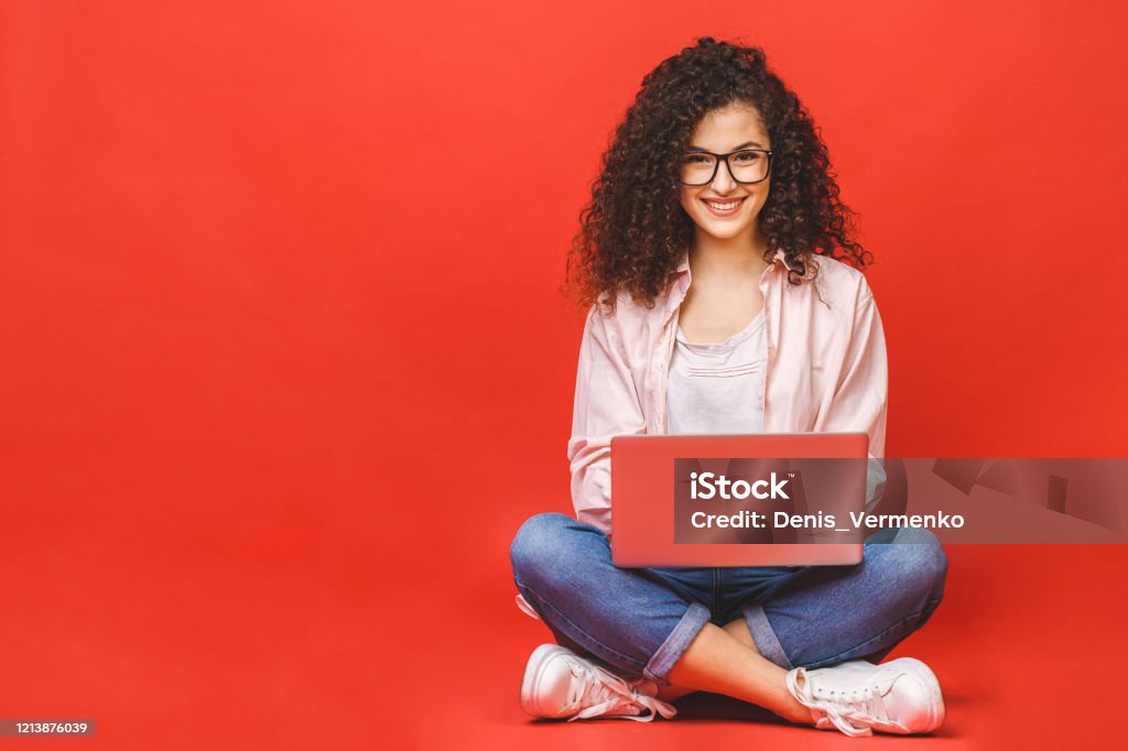 Glad ung lockigt vacker kvinna sitter på golvet med korsade ben och med hjälp av laptop på röd bakgrund. - Royaltyfri Tonåring Bildbanksbilder