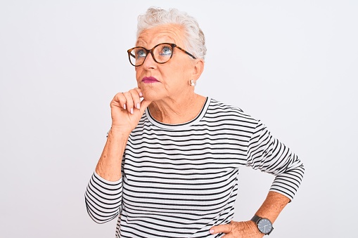 Senior grey-haired woman wearing striped navy t-shirt glasses over isolated white background with hand on chin thinking about question, pensive expression. Smiling with thoughtful face. Doubt concept.