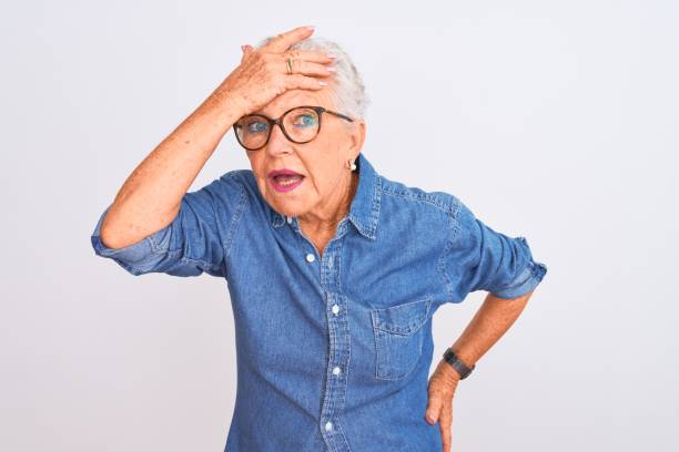 mujer de pelo gris senior con camisa de mezclilla y gafas sobre fondo blanco aislado sorprendido con la mano en la cabeza por error, recuerde el error. olvidé, concepto de mala memoria. - portrait senior adult confusion women fotografías e imágenes de stock