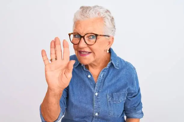 Photo of Senior grey-haired woman wearing denim shirt and glasses over isolated white background Waiving saying hello happy and smiling, friendly welcome gesture