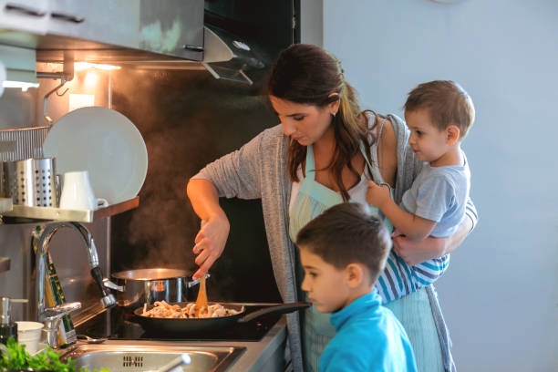 Mother holding toddler and cooking, older son standing by Single mother by the stove with toddler on hip, stirring mushrooms, her older son is standing by family with two children stock pictures, royalty-free photos & images