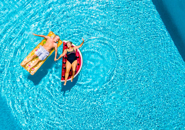 vista vertical de la pareja feliz de personas mayores disfrutando de las vacaciones de verano en la piscina de agua azul con lilos de treny de colores junto con la diversión y el amor - inflatable raft fotos fotografías e imágenes de stock