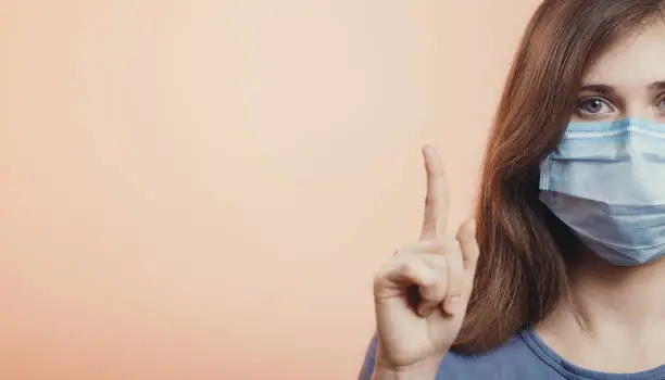 Photo of head of serious young woman raised finger up emphasizing the importance of wearing protective medical mask on colored studio background, concept of desease and quarantine measures