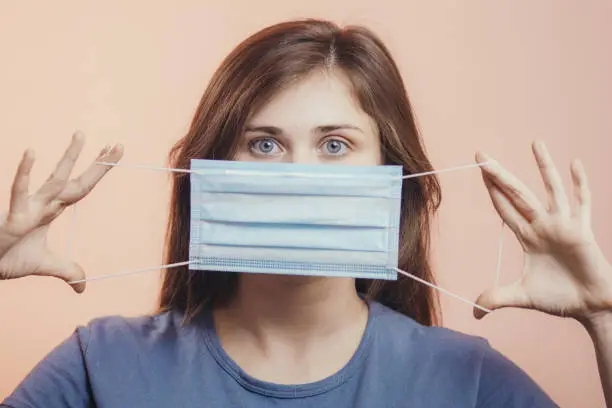 Photo of Portrait of young woman showing opened medical flu mask covering face on colored studio background, girl looking with scared eyes, quarantine measures and panic