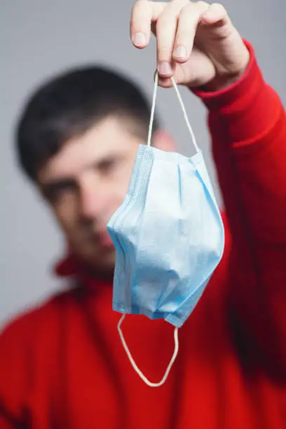 Photo of Portrait of young man holding medical protective mask on studio background, boy take off flu mask, concept of personal responsibility during quarantine, victory over the disease