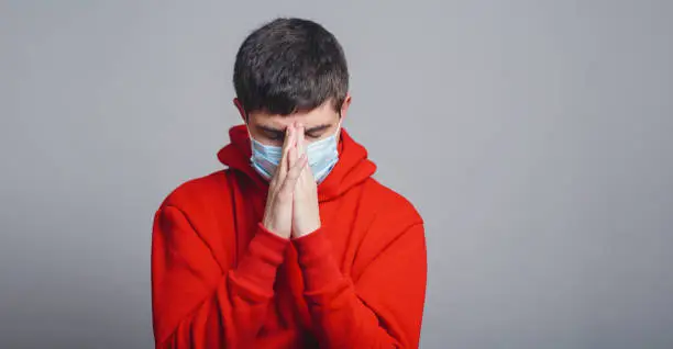 Photo of young man in a protective medical mask on his face bowed his head in sorrow on a gray studio background, concept of health and global epidemic disease,man pray and asks for God's help