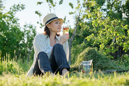 Summertime, woman gardener in hat sitting on freshly cut grass with homemade natural drink mint with strawberries, green garden background