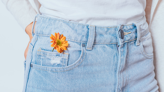Bright orange flower in pocket blue jeans of young slender woman standing on white background, close-up.
