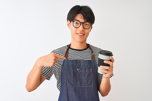Chinese barista man wearing apron and glasses holding coffee over isolated white background with surprise face pointing finger to himself
