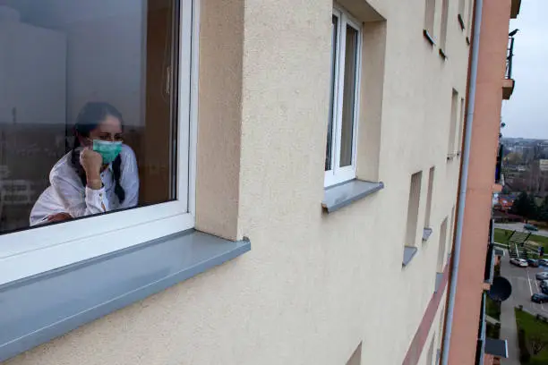 Photo of A 40-year-old woman wearing a protective mask is looking out of the window. Home quarantine for 14 days due to the coronavirus COVID-19 epidemic.