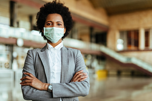 Portrait of African American woman wearing protective mask while standing with arms crossed at the airport during virus epidemic.
