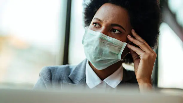 Distraught black businesswoman wearing face mask while being in the office and looking away.
