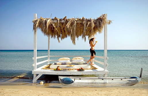 A young girl spends her time on a relaxing white wooden gazebo at a tropical beach. The gazebo is decorated with palm leaves, fruits, pillows and with a comfortable mattress.