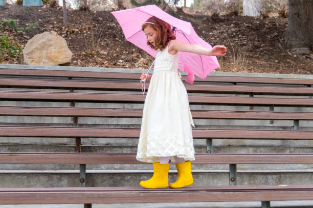 This little girl is holding an umbrella on a rainy spring day.  She is also wearing bright yellow boots and a formal white dress as balances on a bench.