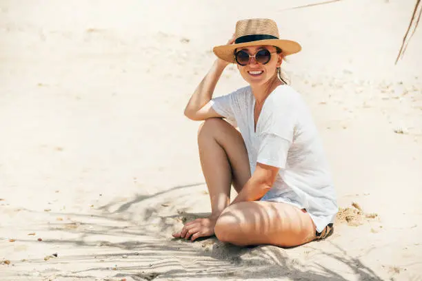 Happy smiling sunbathing woman in straw hat portrait. Palm tree shadows on the sand. Healthy tanning and vacation concept image.