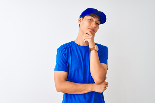 Chinese deliveryman wearing blue t-shirt and cap standing over isolated white background looking confident at the camera smiling with crossed arms and hand raised on chin. Thinking positive.
